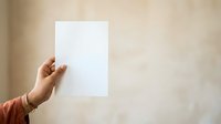 Hand holding blank white paper sheet against neutral beige wall background, suitable for mockup, branding, and presentation designs.
