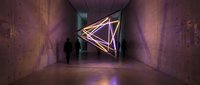 Modern art installation with glowing geometric triangular sculpture in contemporary gallery space. Person silhouette viewing interactive light display in museum corridor.