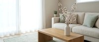 Modern minimalist living room interior with beige sofa, wooden coffee table, white ceramic vase with dried branches, soft natural lighting from window.