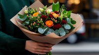 Fresh bouquet with orange roses, red carnations and eucalyptus leaves wrapped in kraft paper, held by person in green sweater. For florist marketing and gift occasions.