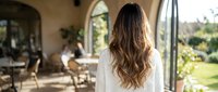 Woman with long wavy brown hair in white sweater standing on terrace overlooking garden patio with outdoor furniture and natural lighting.