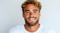 Young Caucasian man with blonde curly hair and beard wearing white t-shirt smiling confidently against light background. Natural genuine expression showing happiness.