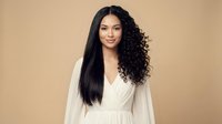 Young mixed-race woman with long curly black hair wearing white dress smiling confidently against beige background for portrait photography.