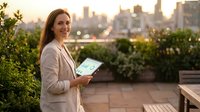 Professional businesswoman holding tablet on urban rooftop terrace at sunset with city skyline background for corporate technology presentations.