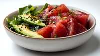 Fresh tuna poke bowl with avocado slices, mixed greens and black sesame seeds served in ceramic bowl on white background, close up view of healthy meal.