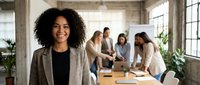 Confident African American businesswoman smiling in modern office with diverse team collaborating in background. Professional workplace leadership concept.