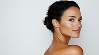 Young mixed race woman with natural makeup and curly hair in updo looking over shoulder against light background, glowing healthy skin and bare shoulders.