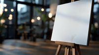 Empty white canvas on wooden easel in modern cafe interior with blurred bokeh lights background, art workshop or gallery atmosphere.