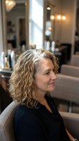 Middle-aged Caucasian woman with curly blonde hair sitting in modern hair salon chair, professional beauty treatment and styling services concept.