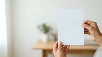 Hands holding blank white paper card in bright interior space with blurred green plant and wooden furniture in background, suitable for mockup, branding, and presentation projects.