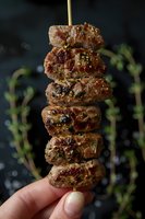 Grilled meat kebab with herbs and spices on wooden skewer held by fingers against dark background. Juicy marinated pieces showcase appetizing char marks.