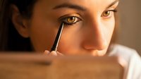 Young Caucasian woman applying black eyeliner with a brush, close-up beauty routine shot with warm natural lighting.