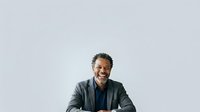 Middle-aged man with dark skin and short dark hair, wearing a charcoal blazer over a navy shirt, smiling warmly at camera against neutral background for corporate and professional use.