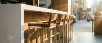 Modern bar counter with wooden stools in bright sunlit cafe or restaurant interior, featuring minimalist design and urban street view through windows.