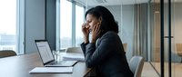 Professional African American businesswoman talking on phone while working on laptop in modern office conference room with city view windows.