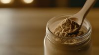 Creamy chocolate protein powder in glass jar with white measuring spoon against warm bokeh background, macro food photography for sports nutrition.