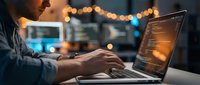 Professional software developer coding on laptop in modern office environment with warm ambient lighting and bokeh background effects.