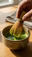 Mature male hand whisking matcha powder in ceramic bowl with bamboo whisk, preparing traditional Japanese green tea ceremony beverage on wooden surface.