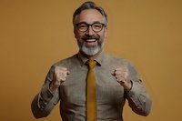 Cheerful mature Caucasian businessman with gray beard and glasses showing success gesture against golden background. Professional portrait expressing achievement.