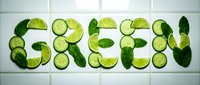 Word GREEN spelled out using fresh cucumber slices, lime wedges, and mint leaves arranged on white tile background, healthy food art concept.