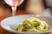 Fresh green pasta ribbons on white plate with cream sauce being poured from ladle in warm kitchen setting for culinary content.