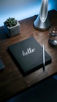 Black notebook with hello lettering on wooden desk with succulent, pen, lamp and glass, modern workspace styled flat lay.