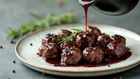Juicy meatballs with rich red wine sauce being poured from pan, garnished with fresh herbs and sesame seeds on rustic plate for gourmet dining experience.