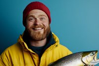 Cheerful bearded fisherman in red beanie and yellow jacket holding fresh caught salmon against blue background, expressing joy and satisfaction with his catch.