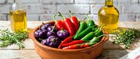 Fresh colorful peppers and herbs in rustic wooden bowl with olive oil bottles on white wooden table for healthy Mediterranean cooking and meal preparation.