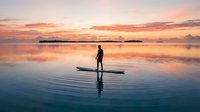 Silhouette of male surfer standing on surfboard in calm ocean waters during golden sunrise with vibrant sky reflection, suitable for travel and lifestyle marketing.
