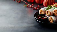 Fresh spring rolls with shrimp and vegetables served on black plate with spicy dipping sauce. Red Asian decor elements and berries create festive mood on dark background.