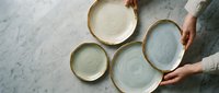 Overhead view of hands holding ceramic plates with natural glazed finishes on marble surface, showcasing tableware and dinnerware styling for home decor and dining concepts.