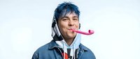 Young man with blue hair, wearing denim jacket and headphones, blowing pink party horn against white background for celebration and fun concepts.
