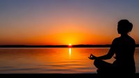Woman meditating in lotus pose by calm lake at golden sunset with peaceful water reflections and serene sky for wellness and mindfulness concepts.
