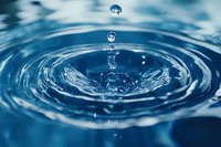 Macro photography captures water drop impact creating concentric ripples and crown splash formation in clear blue water with floating droplets and reflection.