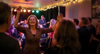 Happy mature woman dancing at evening party celebration with string lights and crowd enjoying festive atmosphere together.