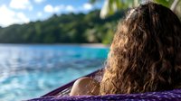 Young woman with curly hair relaxing in purple hammock overlooking tropical beach and turquoise ocean waters, back view capturing serene vacation moment.