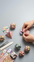 Hands folding colorful patterned origami paper into geometric shapes with crafting tools and decorative papers on gray surface for creative hobby projects.