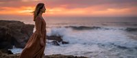 Young woman in flowing brown dress standing on rocky coastline at golden sunset with ocean waves crashing behind her creating romantic silhouette scene.