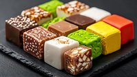 Colorful petit fours and mini dessert cubes arranged in rows on black slate plate, featuring chocolate, vanilla, pistachio and fruit flavored varieties.