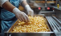 Restaurant kitchen worker in protective gloves preparing fresh cut french fries in metal tray, close-up view of commercial food preparation process.
