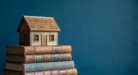 Miniature wooden house model sitting on stack of vintage books against blue background representing real estate education and property investment learning.