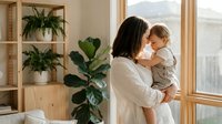 Loving mother holding adorable toddler in bright modern home with plants and natural light streaming through large windows creating warm family moment.