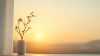 Minimalist white ceramic vase with delicate branch silhouette against warm golden sunset window view creating peaceful home interior atmosphere.