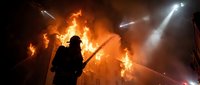 Firefighter silhouette battling intense house fire with dramatic flames and smoke illuminated by emergency spotlights during nighttime rescue operation.