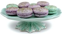 Pastel purple and mint green macarons with gold glitter sprinkles arranged on vintage-style mint ceramic cake stand with leaf pattern base, isolated on white background.