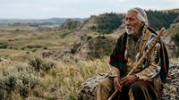 Senior man with white beard and glasses, wearing earth-tone jacket and beaded necklace, sitting on rock overlooking vast valley landscape with rolling hills and sparse vegetation. Native American or indigenous heritage appearance. Contemplative outdoor portrait for editorial, cultural, lifestyle, and travel use.