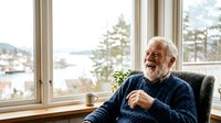 Elderly man with white beard and glasses sitting by large window overlooking water and trees, wearing dark sweater, smiling during conversation or interview setting.