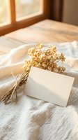 Blank white card leaning against dried golden flowers on neutral linen fabric near sunlit window, suitable for wedding invitations, greeting cards, and stationery design mockups.