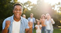 Man of African descent, wearing light blue shirt over white t-shirt, showing excited expression with fists raised in celebration gesture outdoors with blurred group of people in background during golden hour sunlight, conveying joy and success for motivational, lifestyle, and community themes.